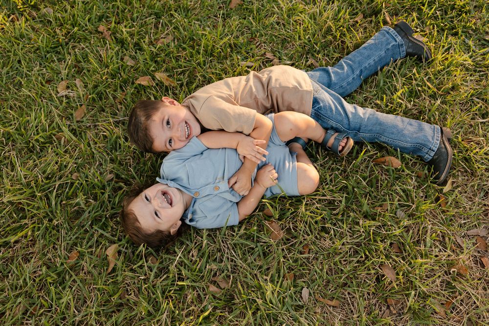 Two young boys laughing and playing together while lying on green grass outdoors.