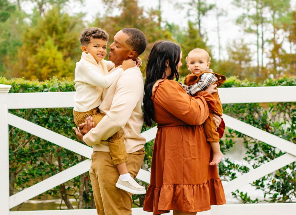 A family dressed in autumn colors shares a tender moment together by a white fence on a sunny day.