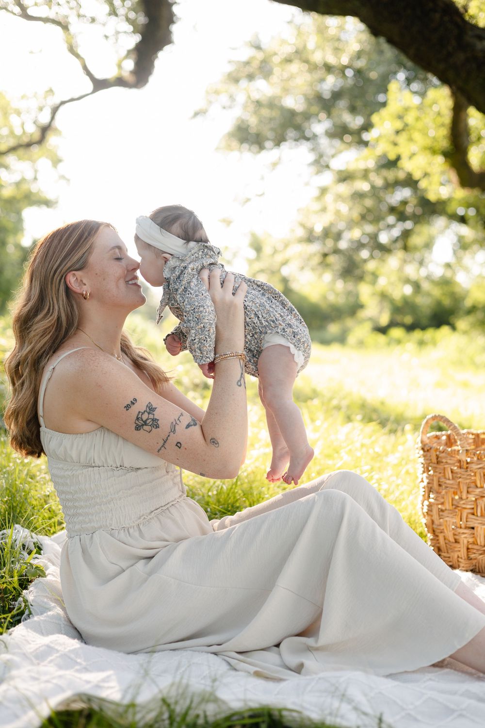 A smiling mother sits on a blanket outdoors, lifting her baby girl in a floral outfit, enjoying a sunny picnic.