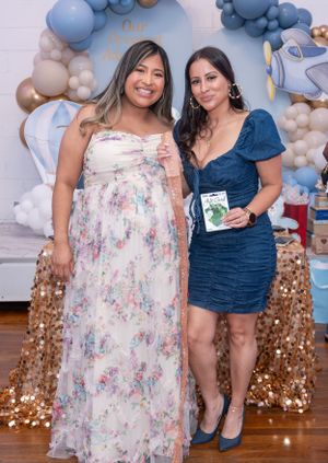 Baby shower celebration with two women posing in front of blue and beige balloon arch decor.