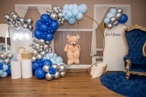 Baby shower decoration with blue and silver balloons, teddy bear, and royal blue throne chair.