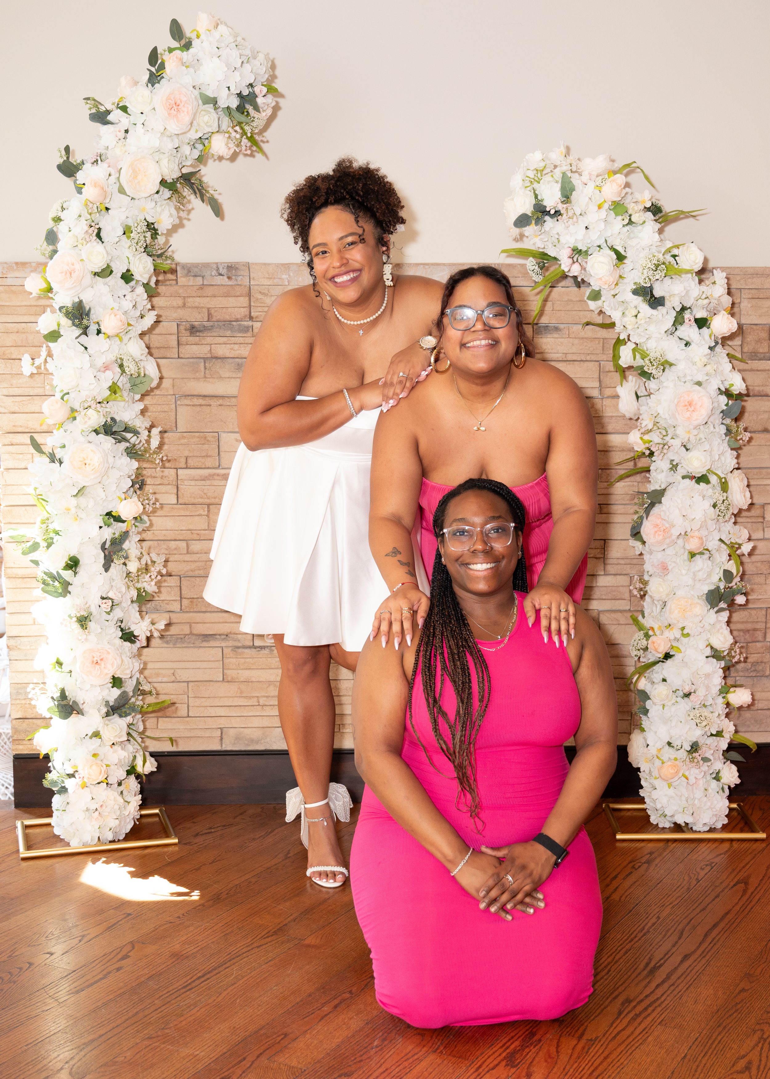 Friends pose together in front of a floral arch with white flowers against a brick wall.