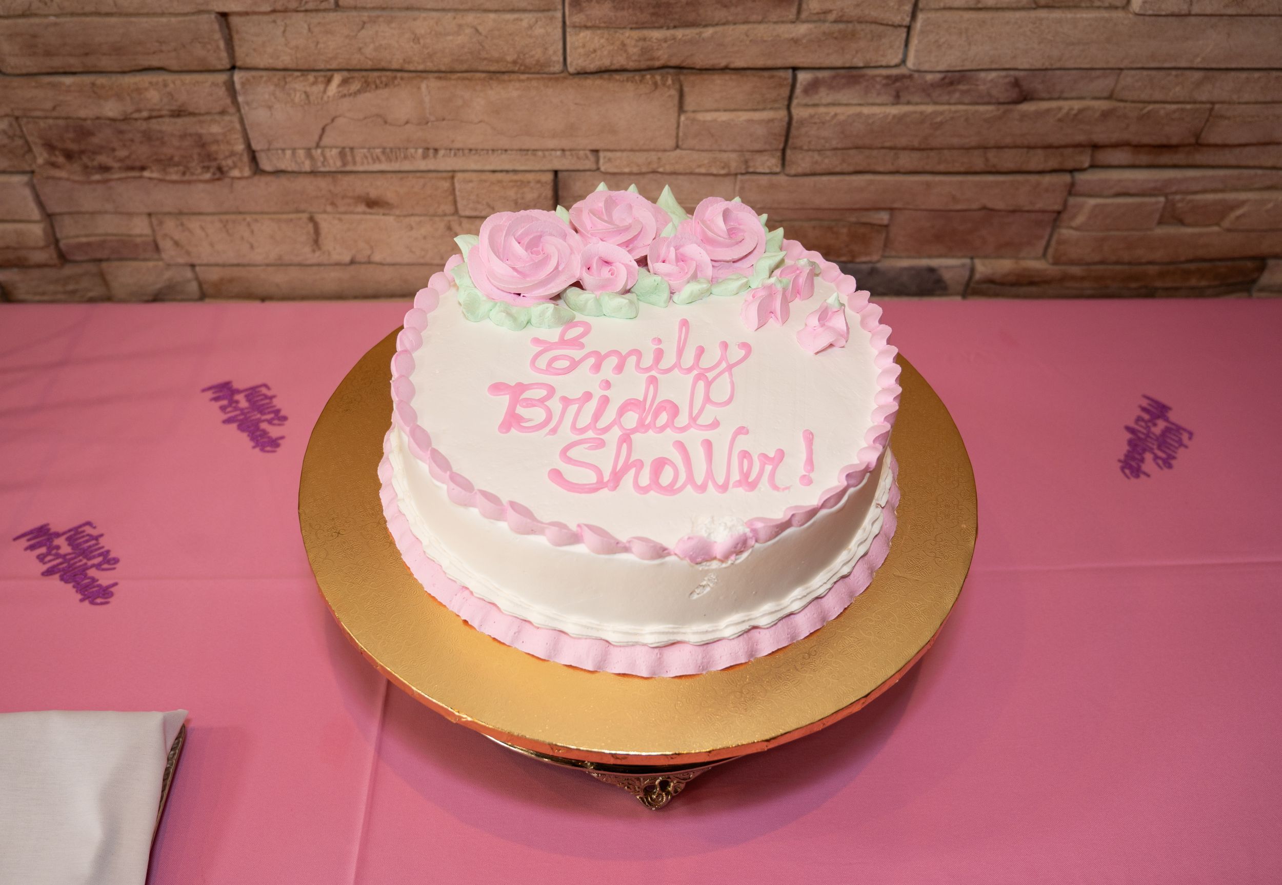 A round white birthday cake with pink frosting roses and "Happy Birthday" text sits on a gold cake stand against a pink tablecloth.