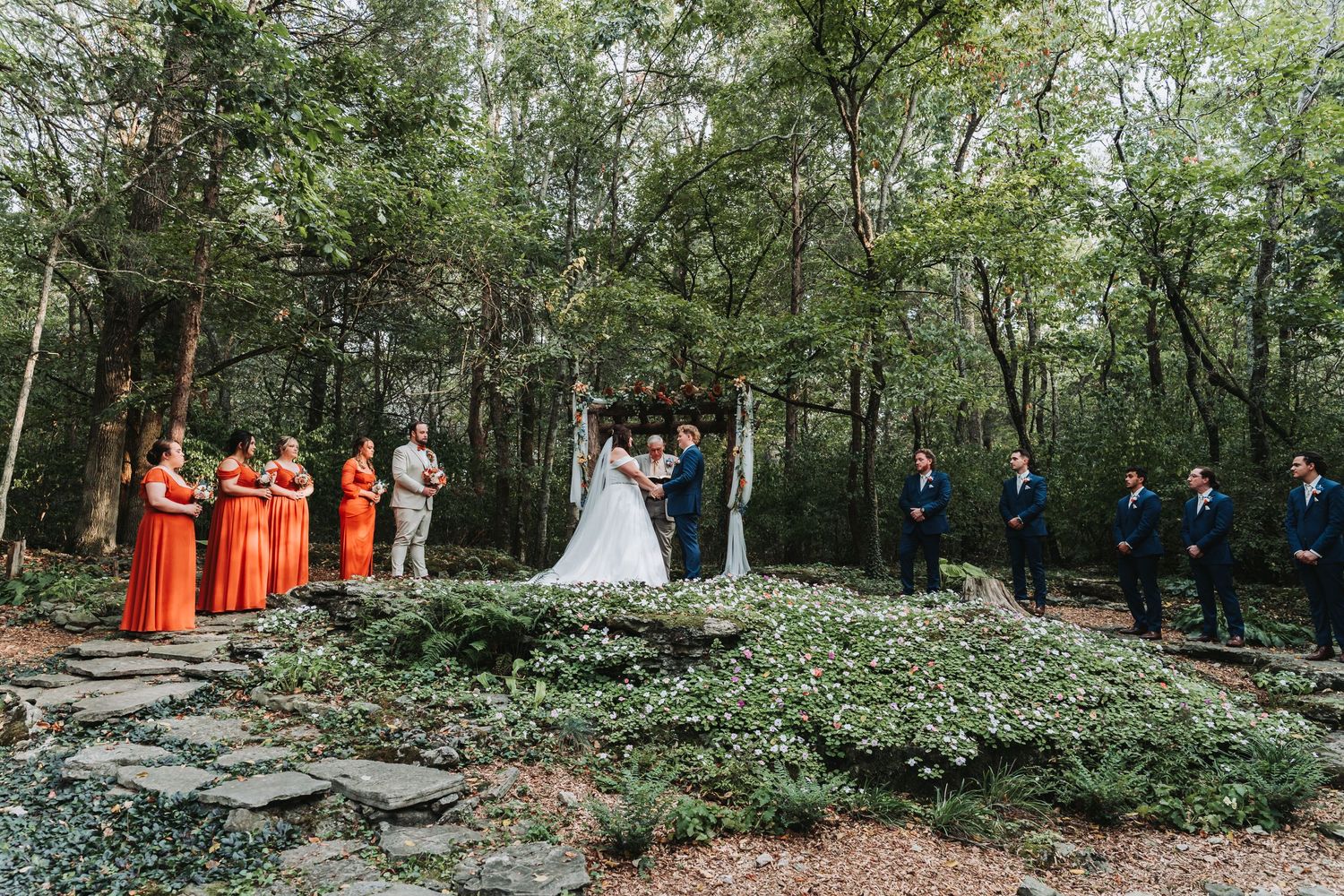 Wedding ceremony takes place in a secluded wooded area with stone platform surrounded by bridesmaids in orange dresses.