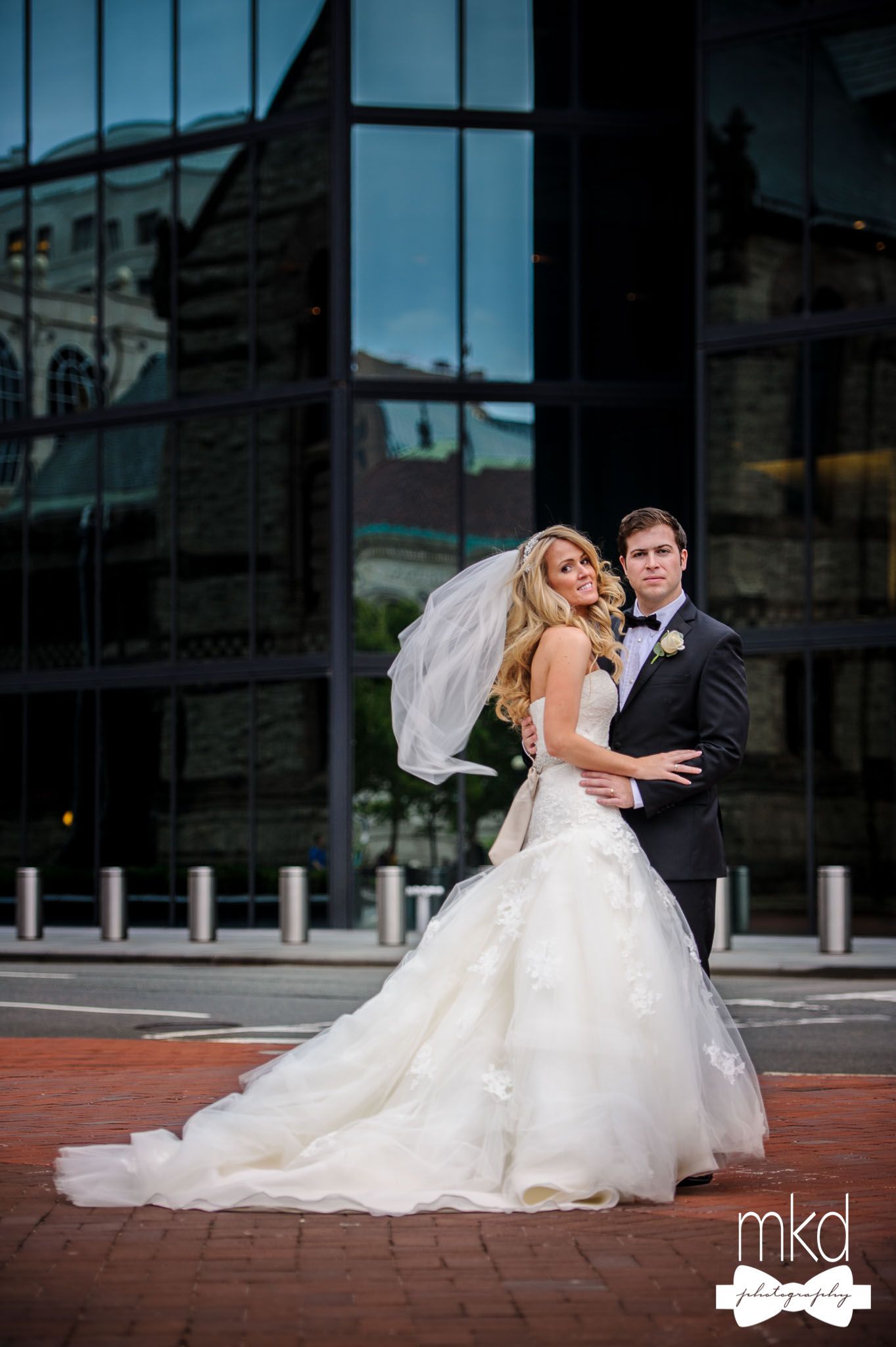 Elegant wedding couple poses against modern glass building exterior in urban setting.