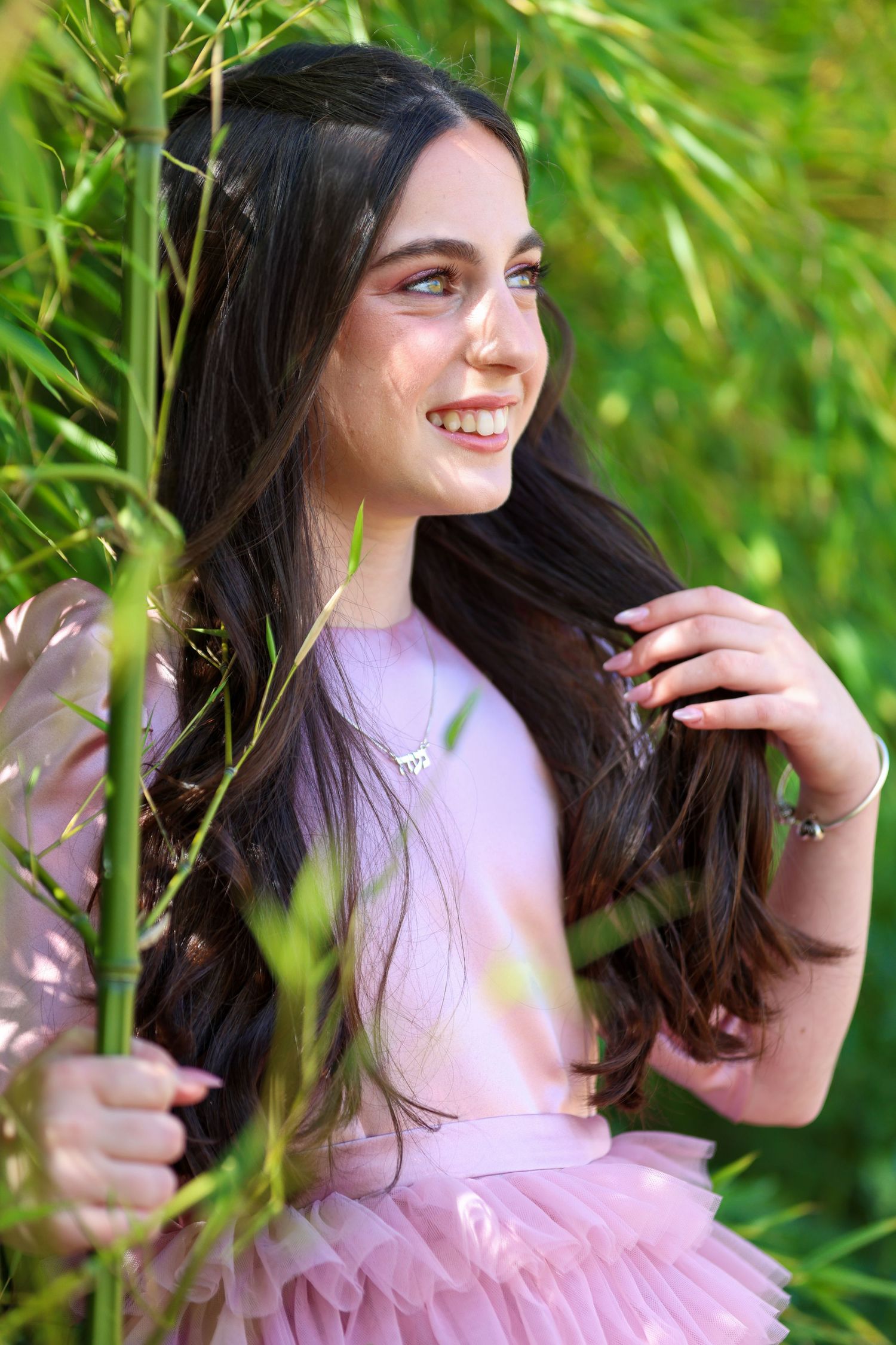 Smiling teenage girl with long dark hair in pink dress among green bamboo.