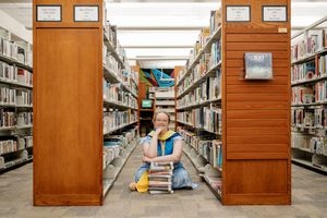 A smiling person sits on a library floor surrounded by tall bookshelves, holding a stack of books.