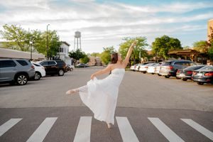 Ballerina in white dress dancing gracefully on a crosswalk in an urban street setting with cars and trees.