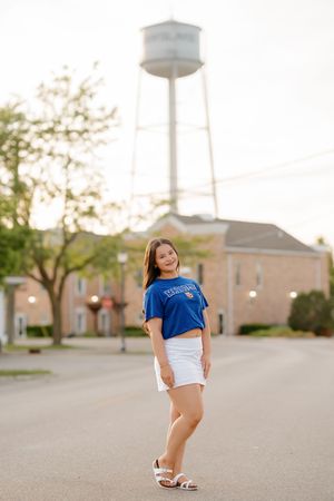 Young woman in blue university shirt and white skirt smiling on a street with a water tower backdrop.