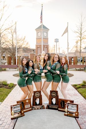 Six young women in green outfits pose with trophies and awards in front of a clock tower at sunset.
