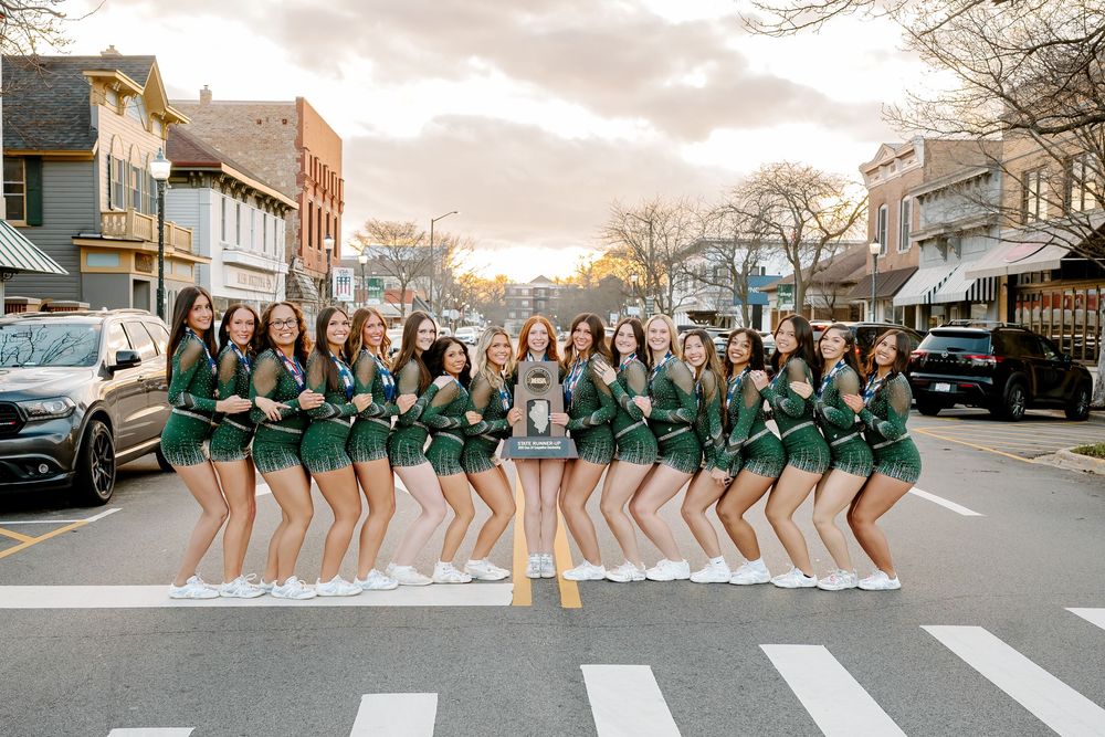 A cheerleading squad in green uniforms poses on a crosswalk in a small town at sunset.