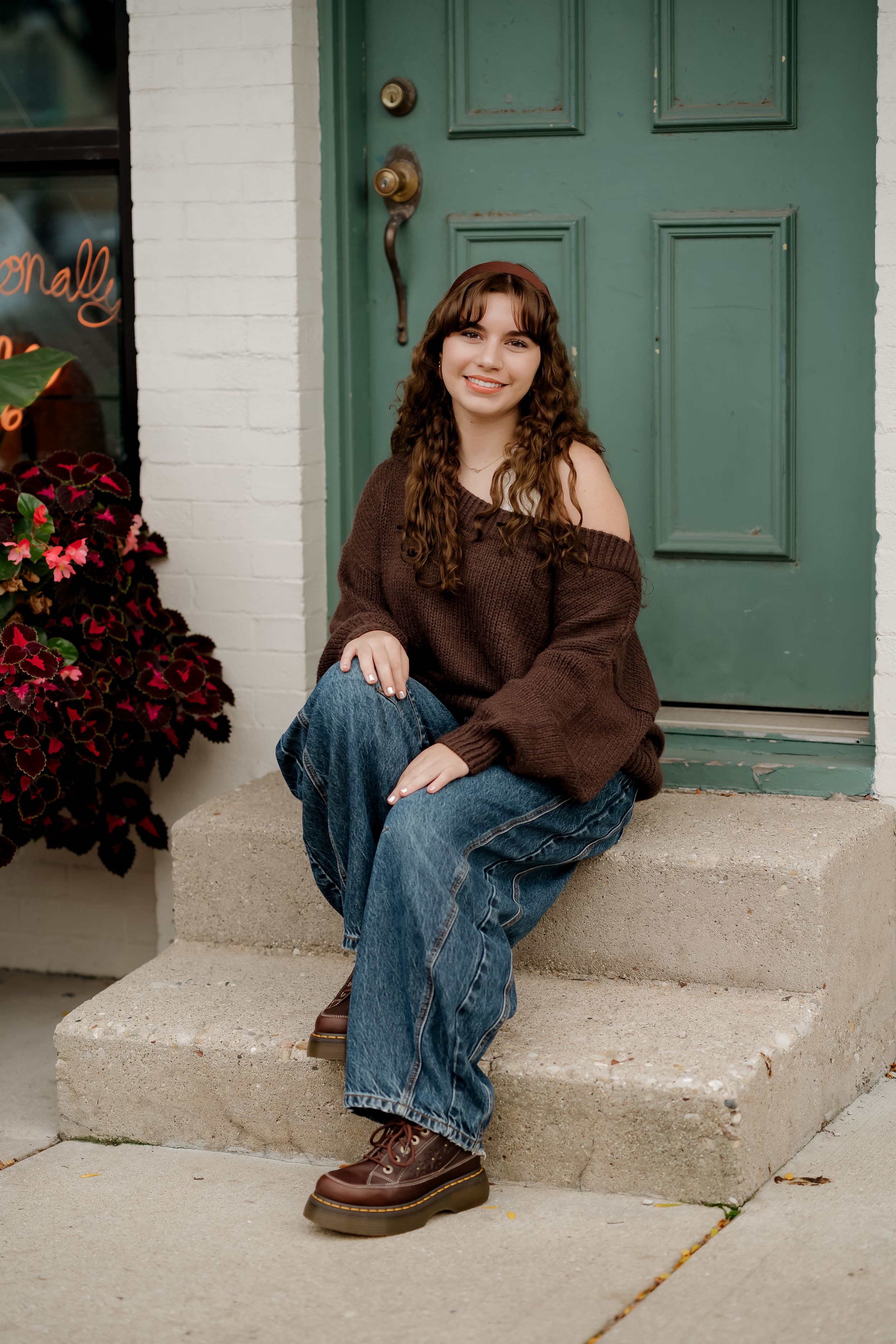 Young woman in brown off-shoulder sweater and jeans sits smiling on concrete steps by a teal door.