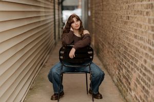 Young woman in brown top and jeans sits on a chair in a narrow brick alley for a senior portrait photo.