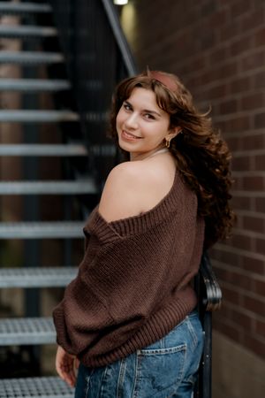 Young woman in brown off-shoulder sweater and jeans smiling over shoulder near brick wall and metal stairs.