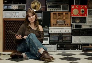 Young woman sitting on checkered floor holding tablet, surrounded by vintage stereos and radios stacked against wall.