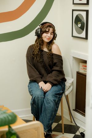 Young woman wearing headphones and brown off-shoulder sweater sitting on stool in music-themed room.