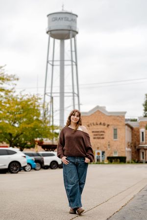 Young woman in brown sweater and jeans poses on street near Grayslake water tower and Village Municipal History Museum.