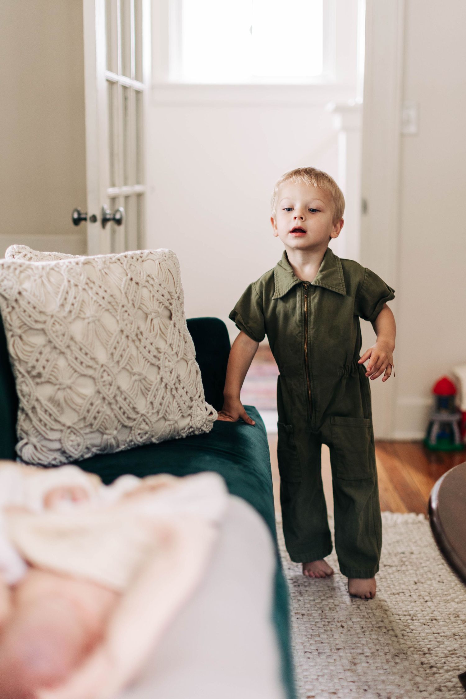 Small child in olive green jumpsuit stands next to couch with textured throw pillows.
