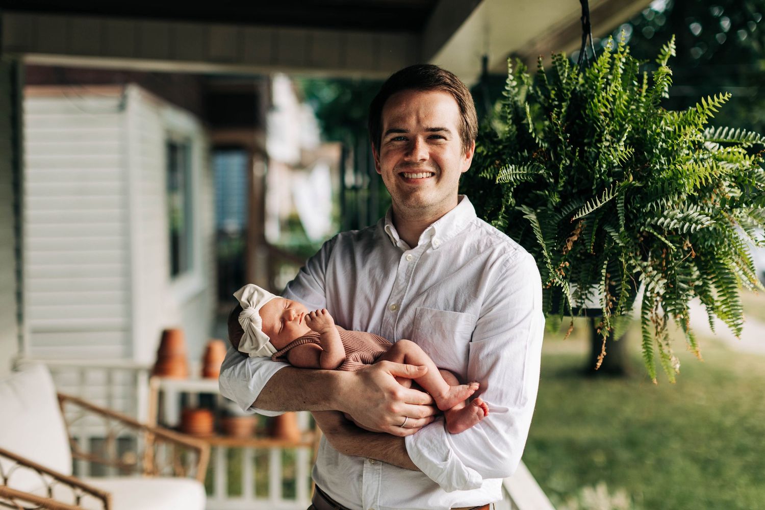 Joyful parent holding newborn baby outdoors near lush hanging plants.