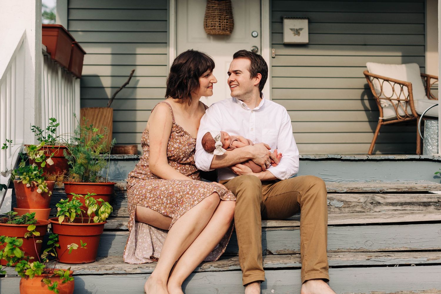 Couple sitting on porch steps with newborn baby surrounded by potted flowers.