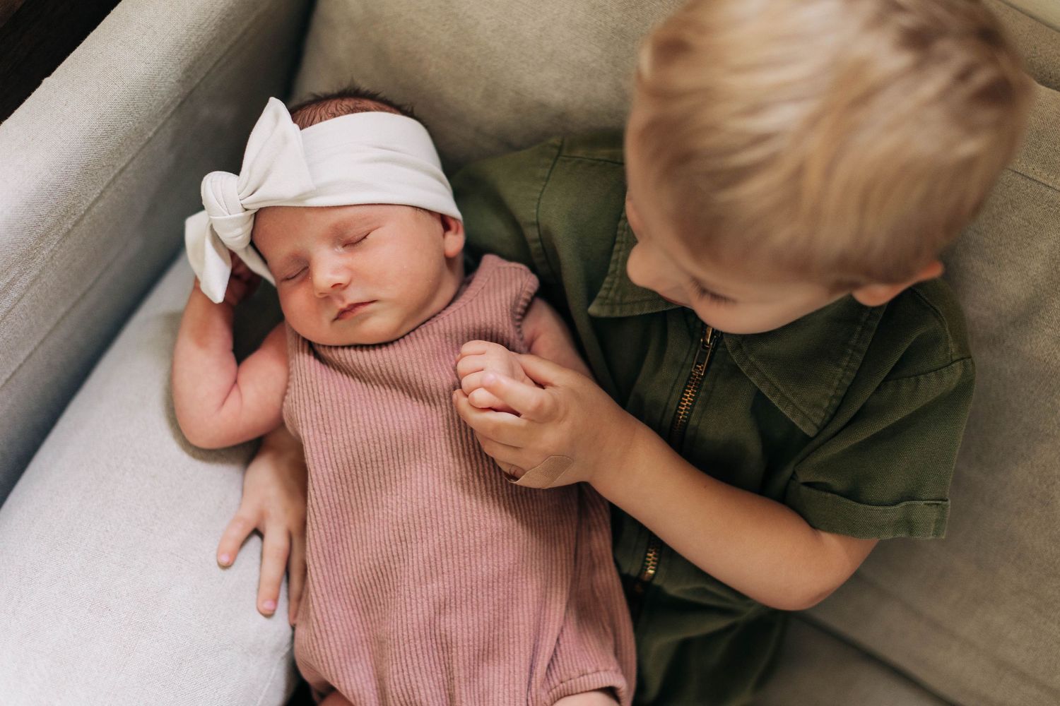 Toddler in green outfit gently holds newborn's hand while sitting on beige couch.