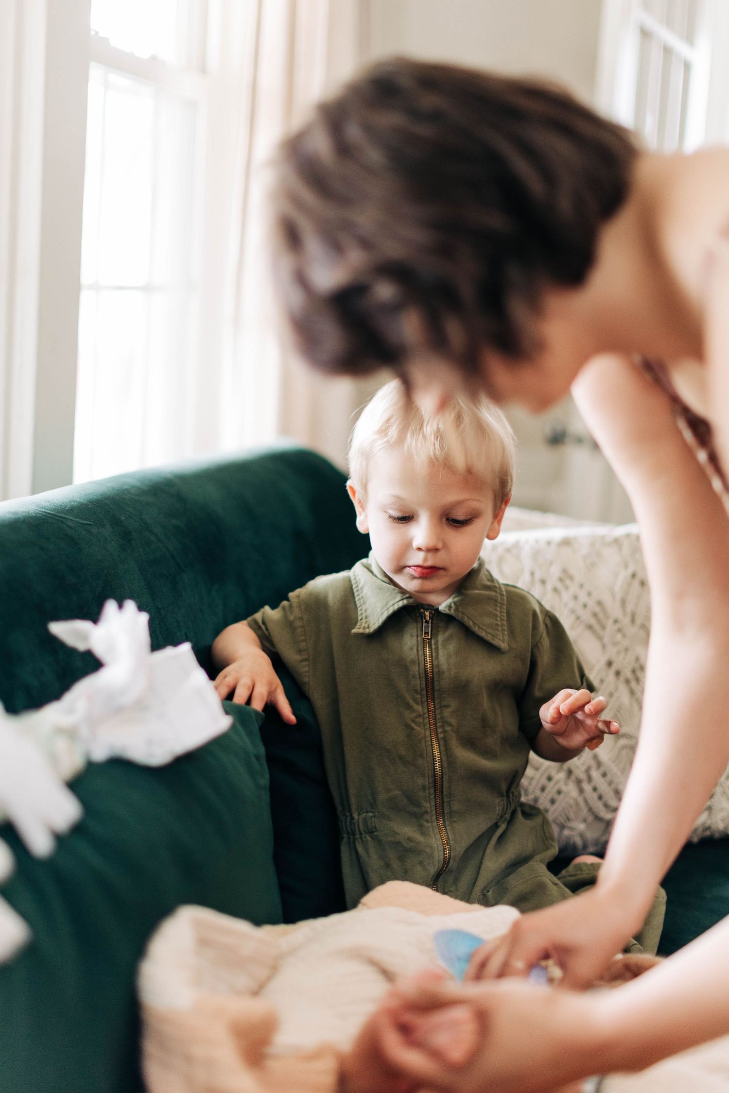 Child in olive green outfit stands by emerald couch while hands offer a white plush toy.