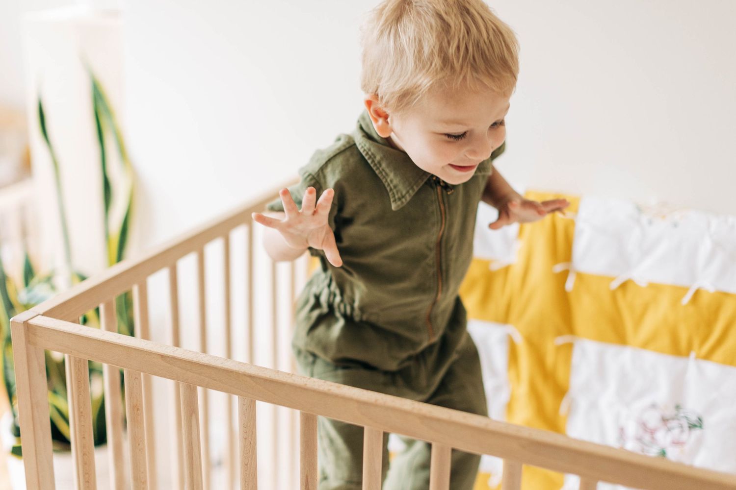 Toddler in olive green outfit standing inside wooden crib with yellow bedding.