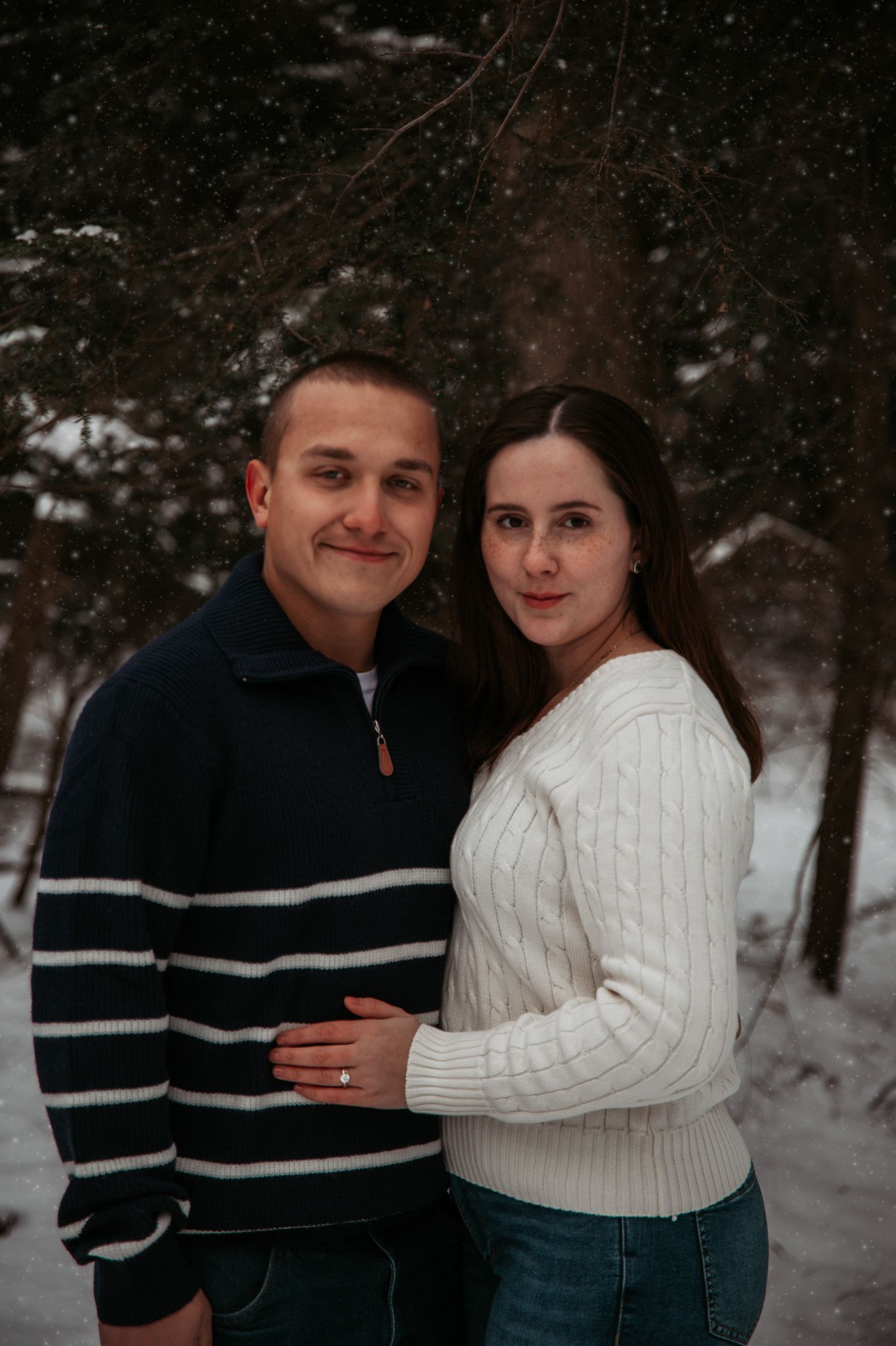 A couple poses together in a snowy forest scene wearing coordinating sweaters.