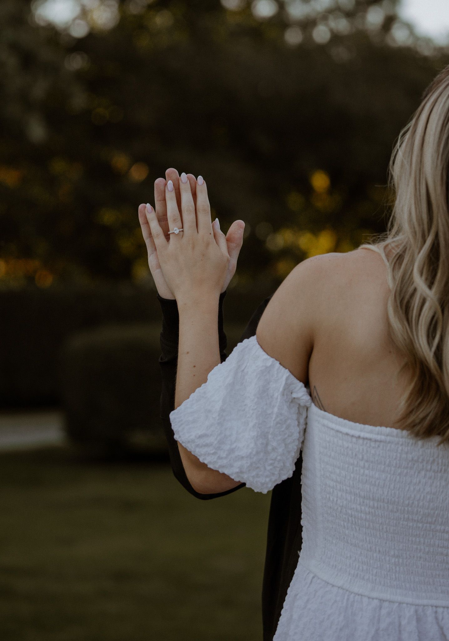 Close-up of a raised hand wearing an engagement ring with a white off-shoulder dress in an outdoor setting.