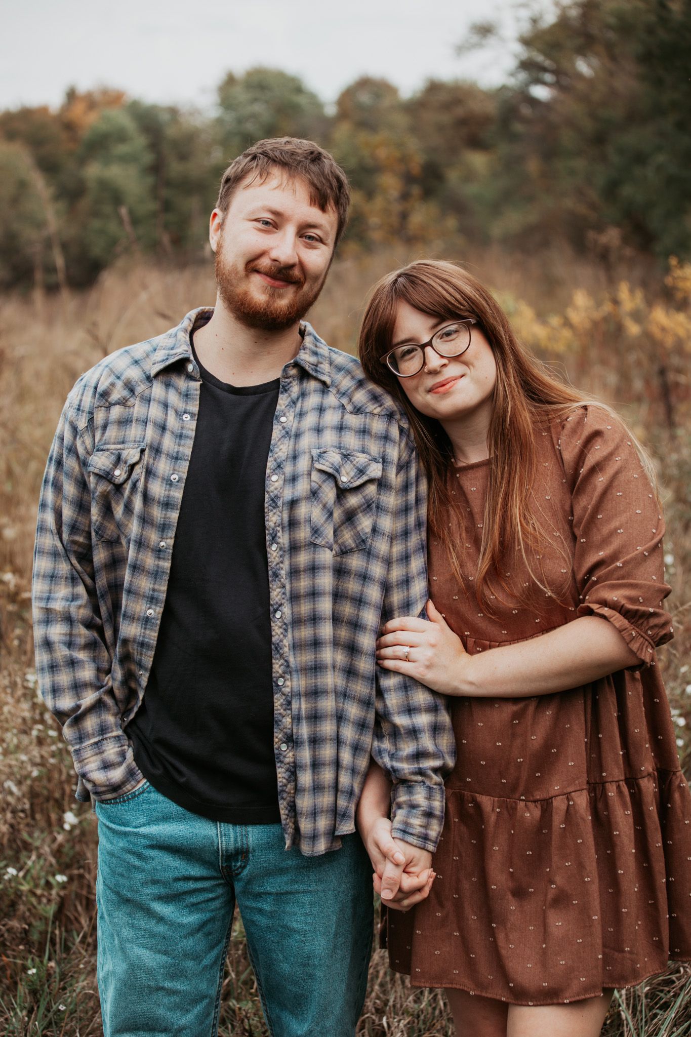 A couple stands together in a fall field wearing casual clothing and sharing a tender moment.
