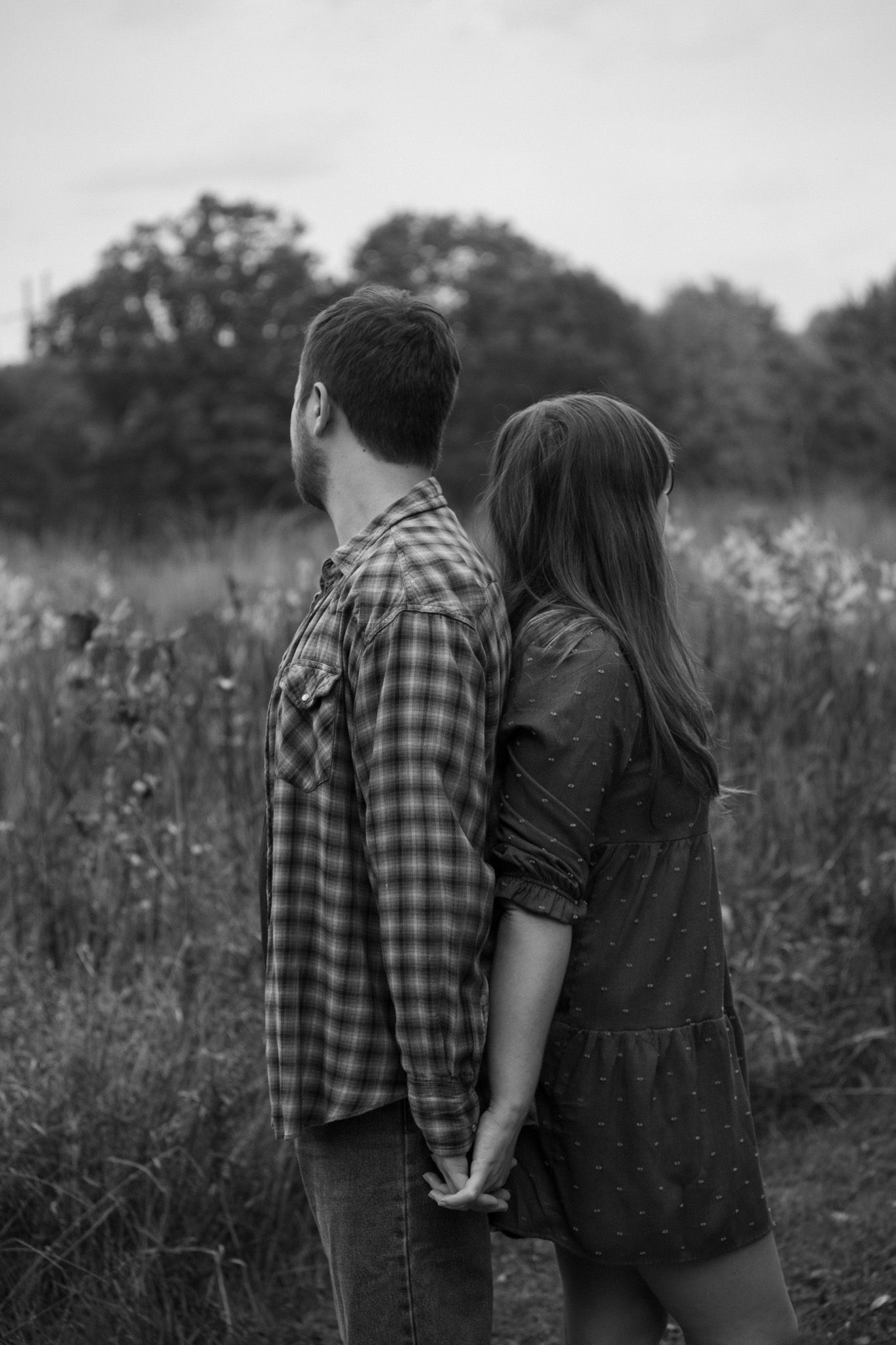 A couple stands back to back holding hands in a field during golden hour.