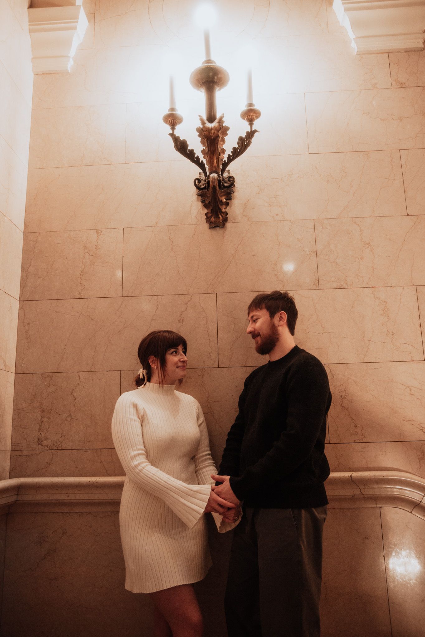Couple sharing intimate moment under ornate wall sconce in marble hallway.