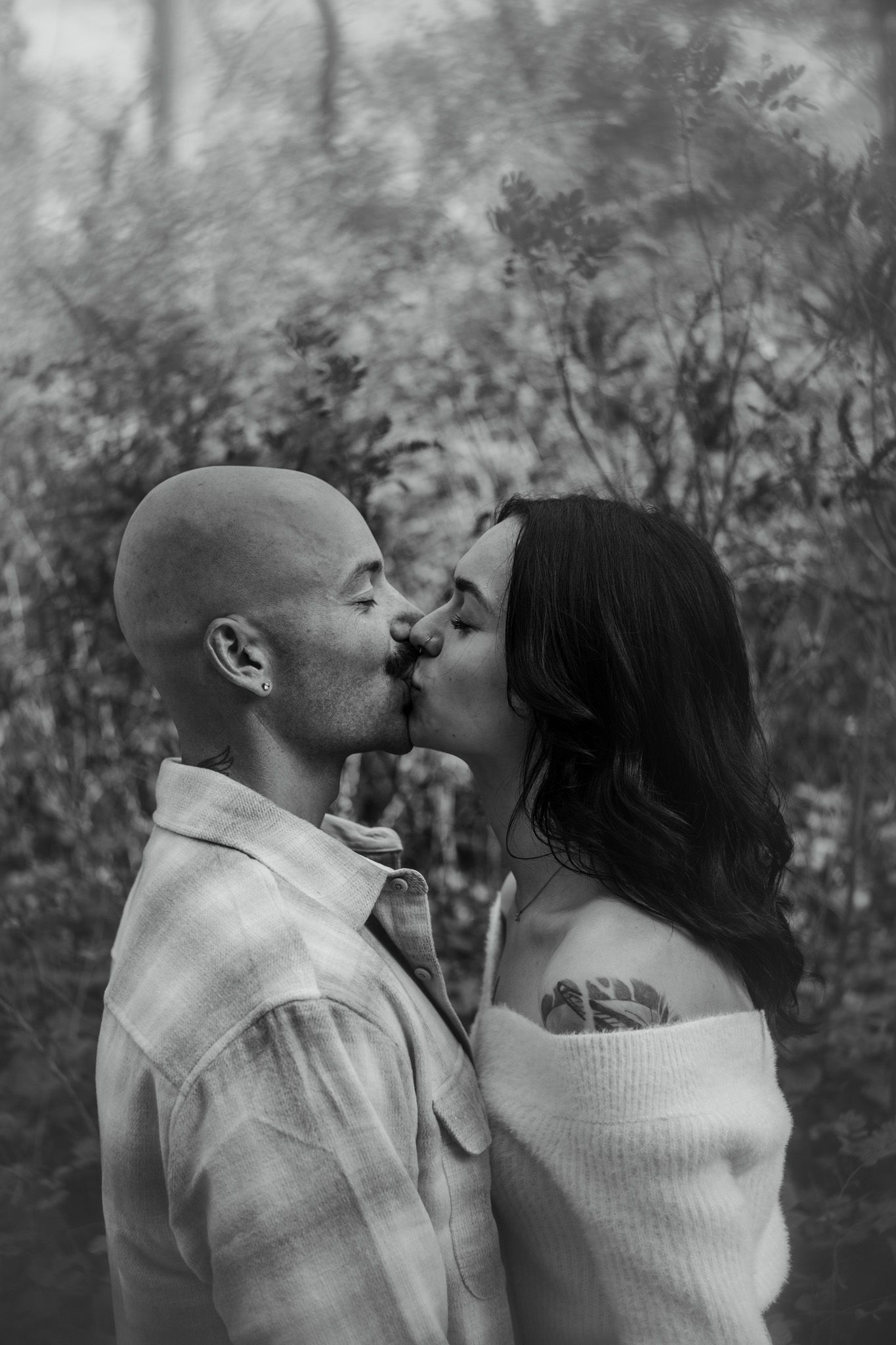A couple in white sweaters shares tender kisses in a black and white outdoor portrait.