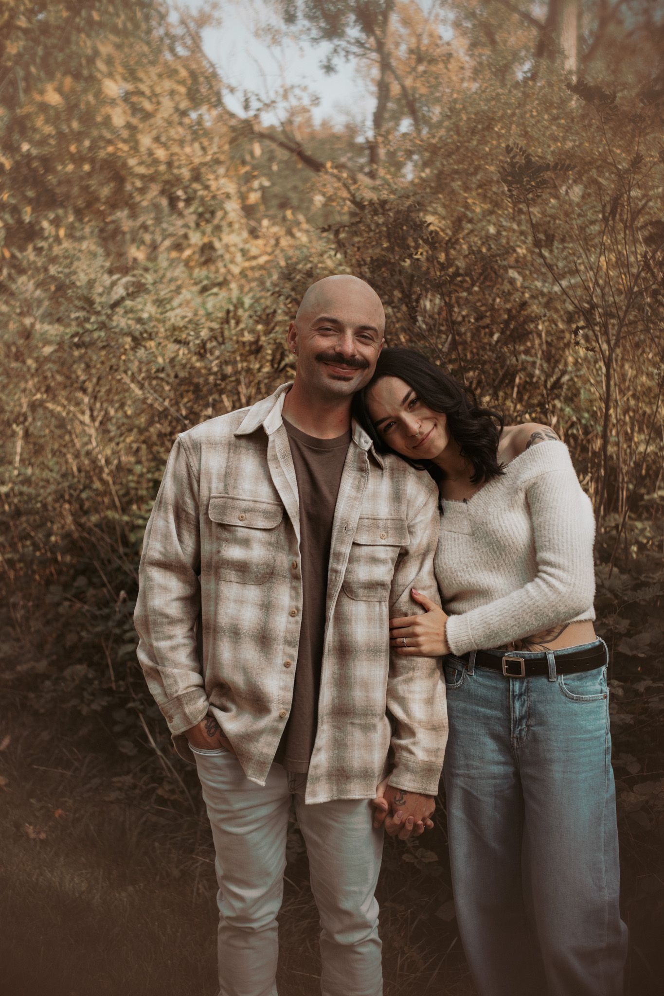 A couple in matching plaid jackets embraces in a warm autumn setting.