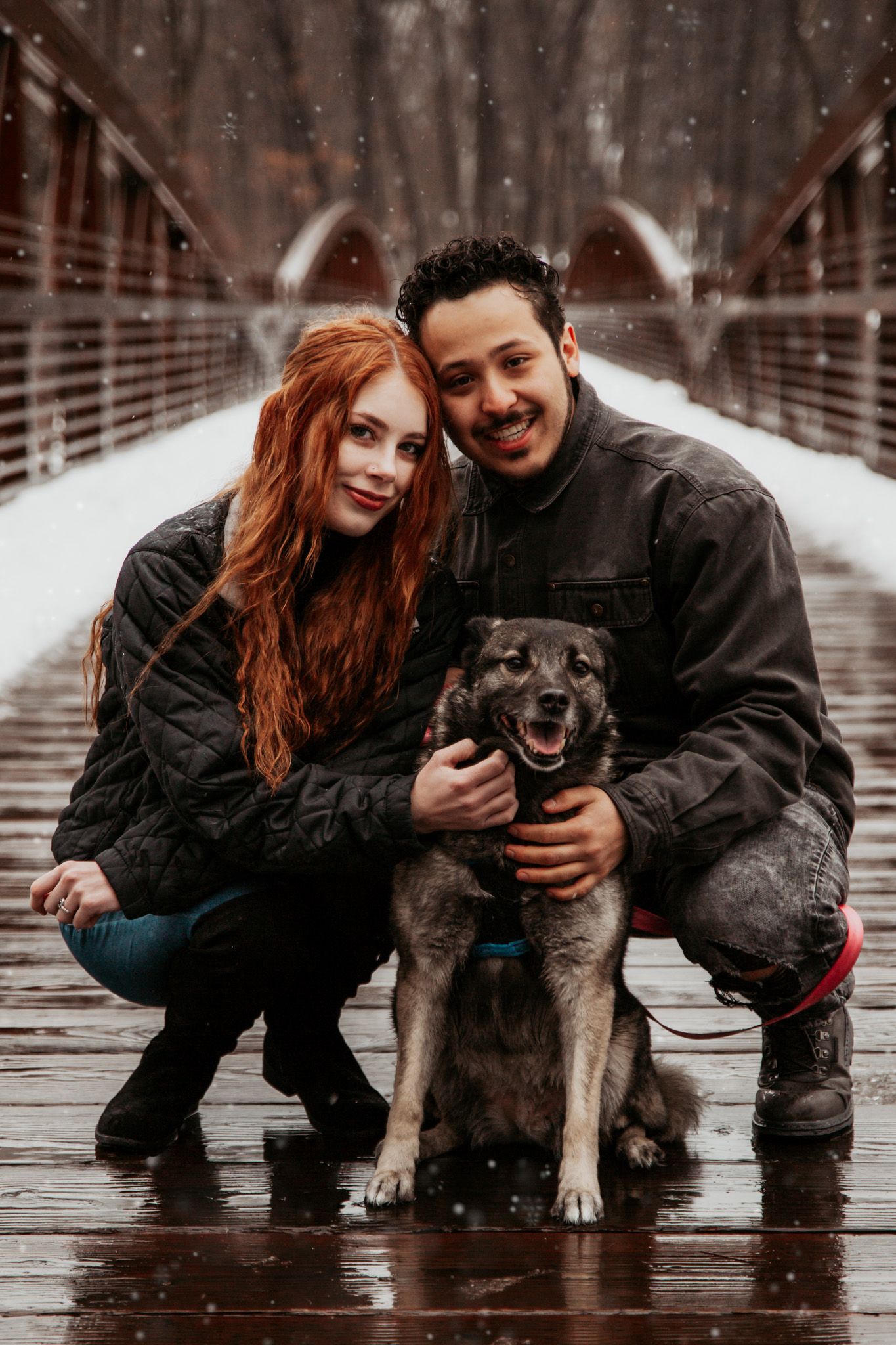 Happy couple with their dog on wooden bridge during snowy winter day.