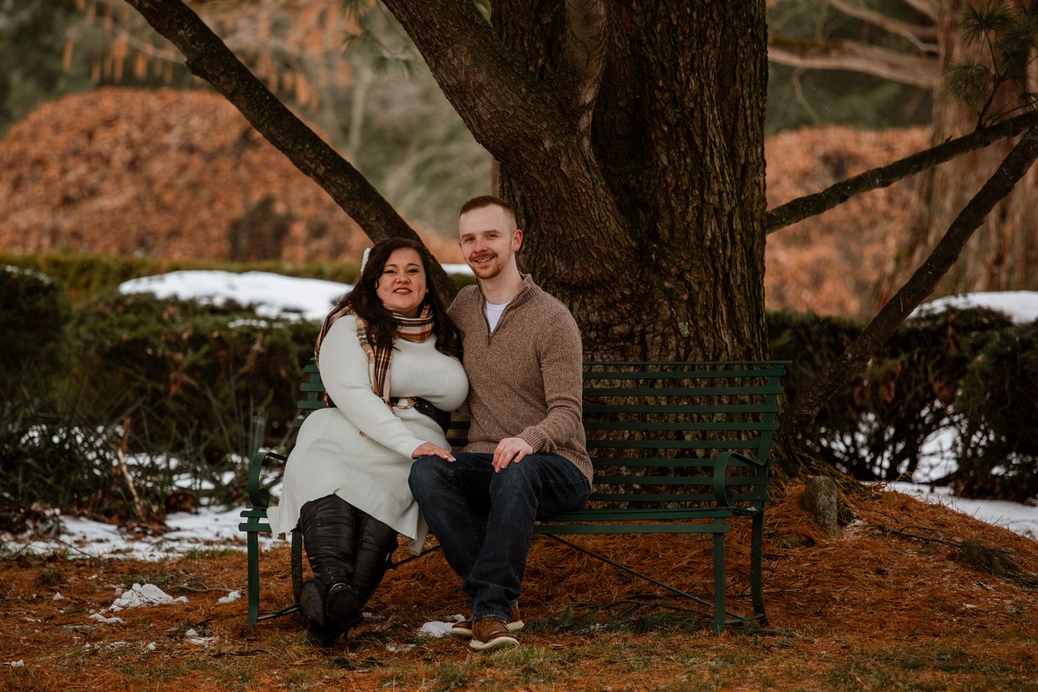 A couple embraces in a romantic autumn setting with fallen leaves and trees in the background.