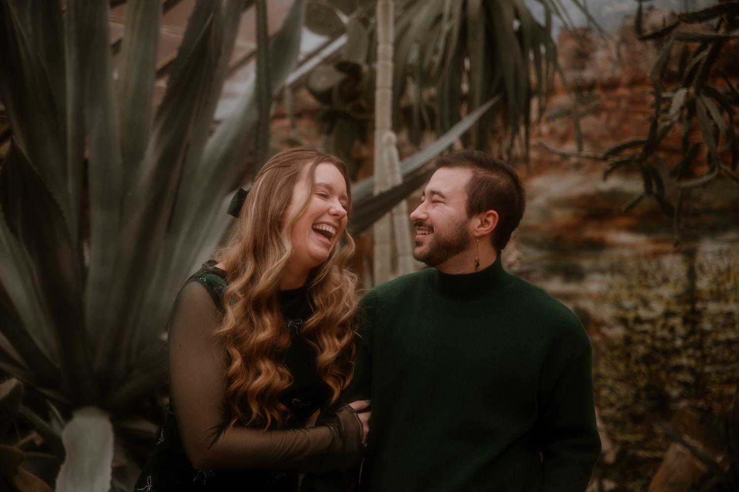 A couple shares a joyful moment together surrounded by greenery in warm lighting.