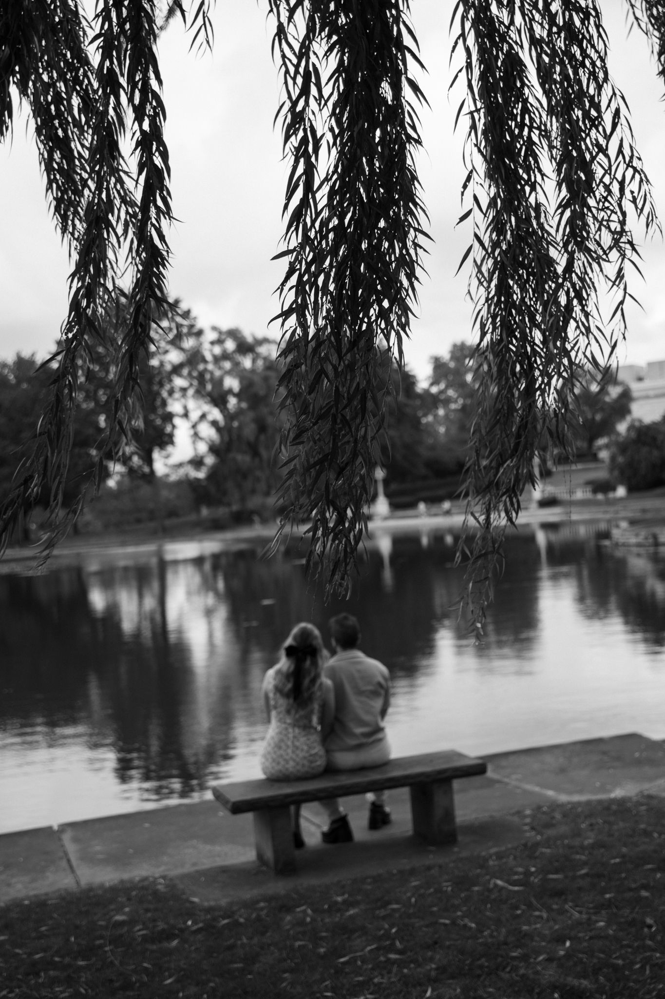 A couple sits on a bench by a lake under weeping willow trees in black and white.