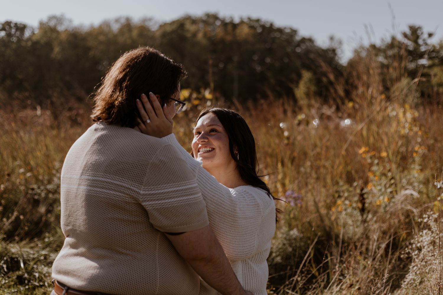 A couple embraces in a sunlit field surrounded by autumn foliage.