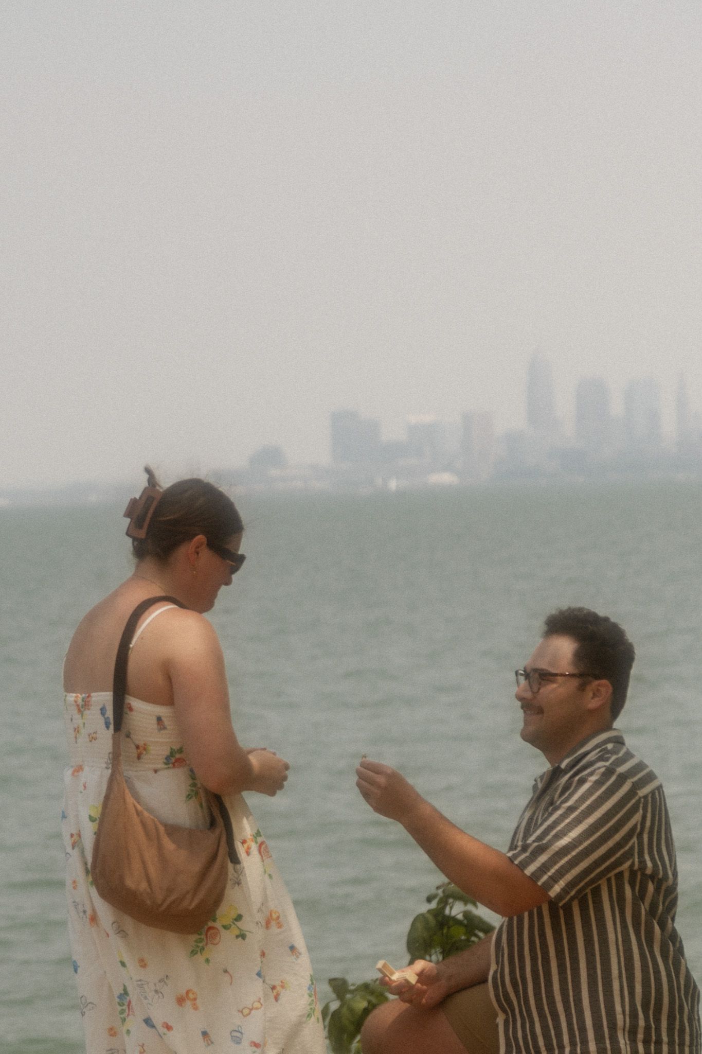 A couple stands by the waterfront with a city skyline visible in the distance.