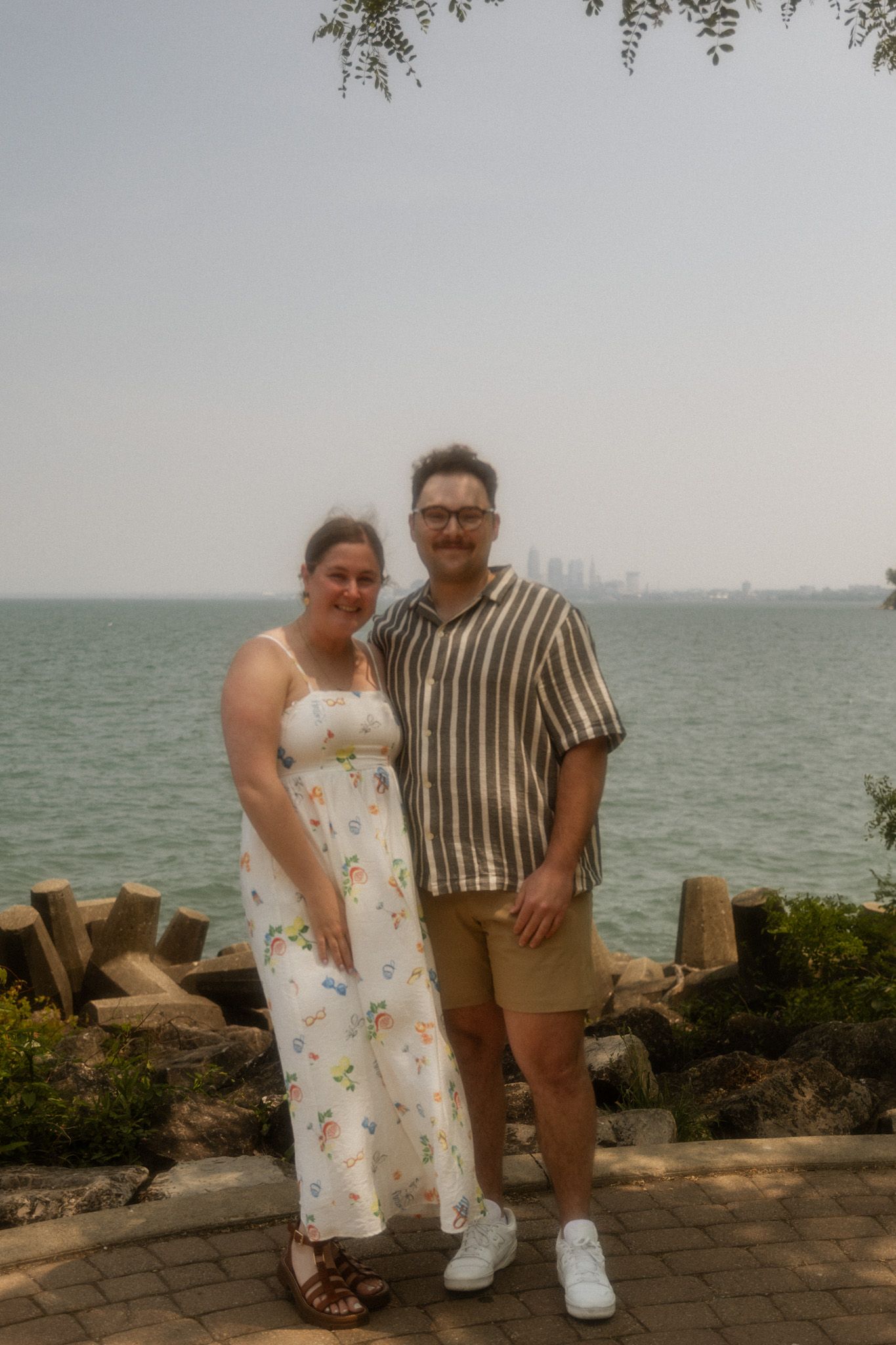 Two people pose together on a stone path near the water with a cityscape in the background.