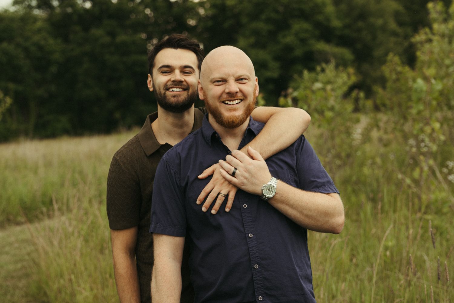 Two smiling men embrace outdoors in a green field, one resting his chin on the other's shoulder.
