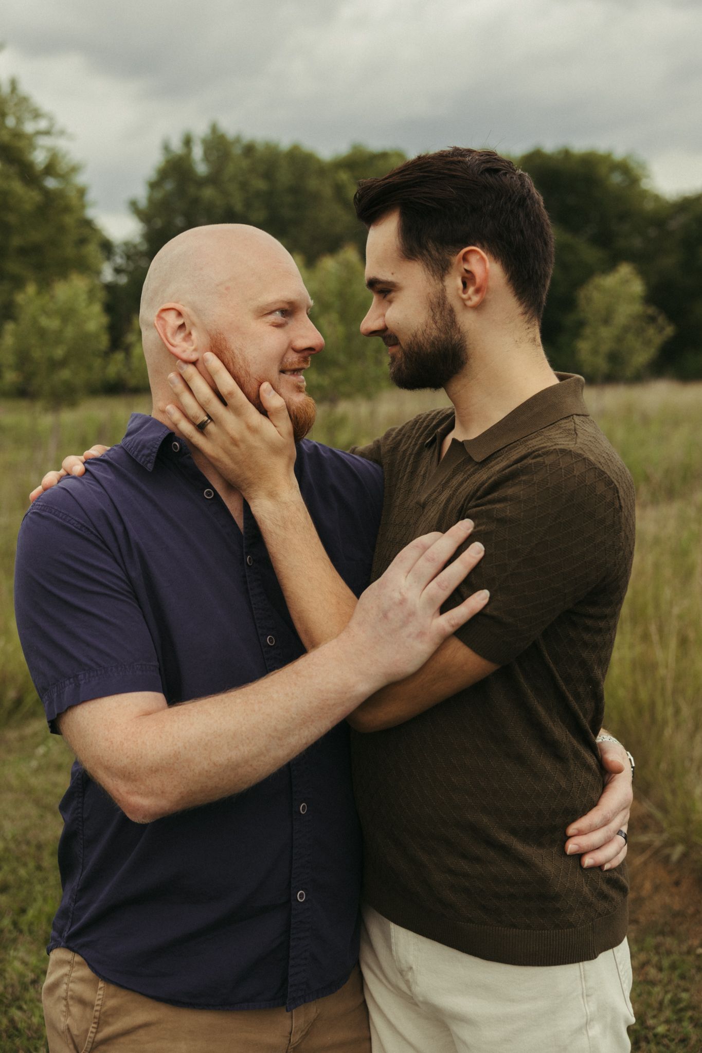 Two men embrace tenderly in a green field, gazing into each other's eyes on a cloudy day.