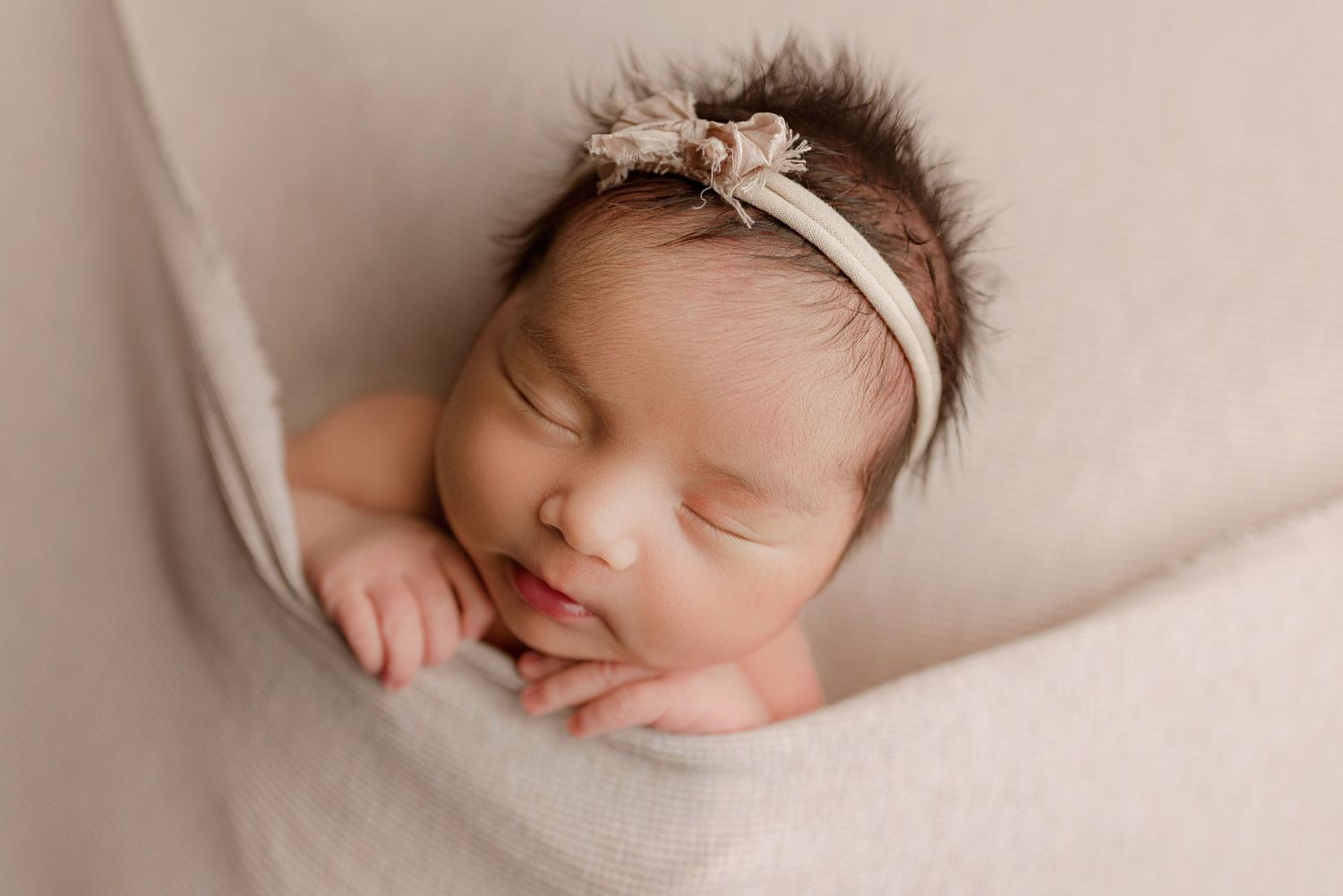 A peaceful newborn baby girl with dark hair sleeps wearing a floral headband on a soft beige blanket.