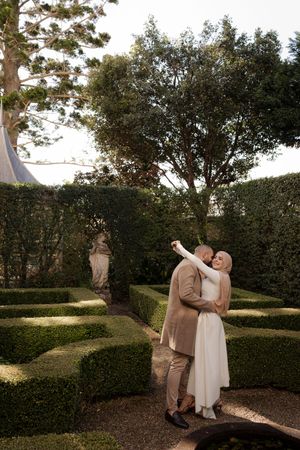 Couple in elegant attire dance joyfully in a formal manicured garden with sculpted hedges and statues.