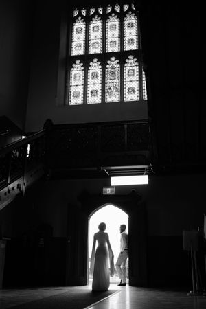 Black and white silhouette of bride and groom holding hands beneath stained glass window in church.