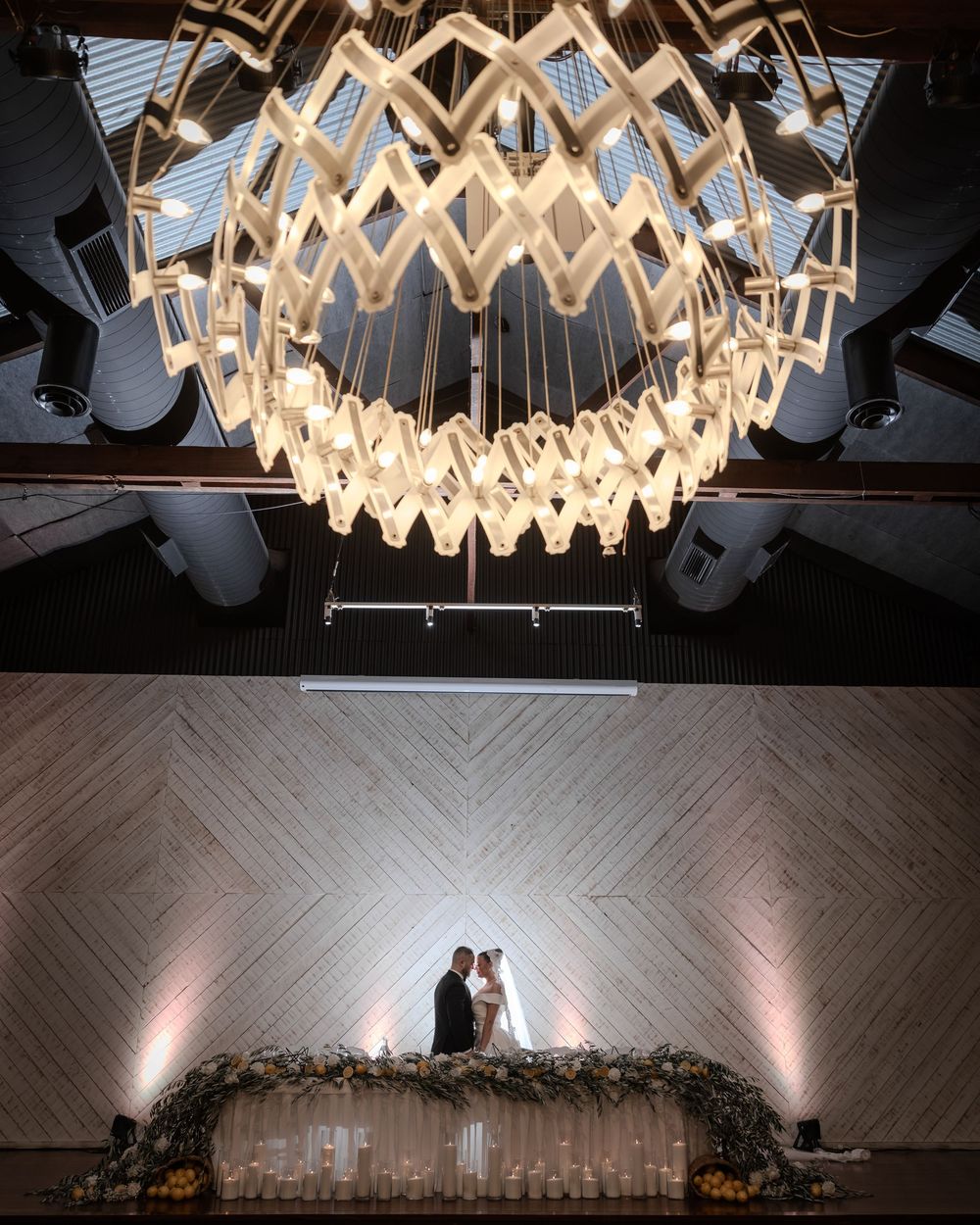 Couple shares intimate moment beneath an ornate geometric chandelier above a floral arch with candles.