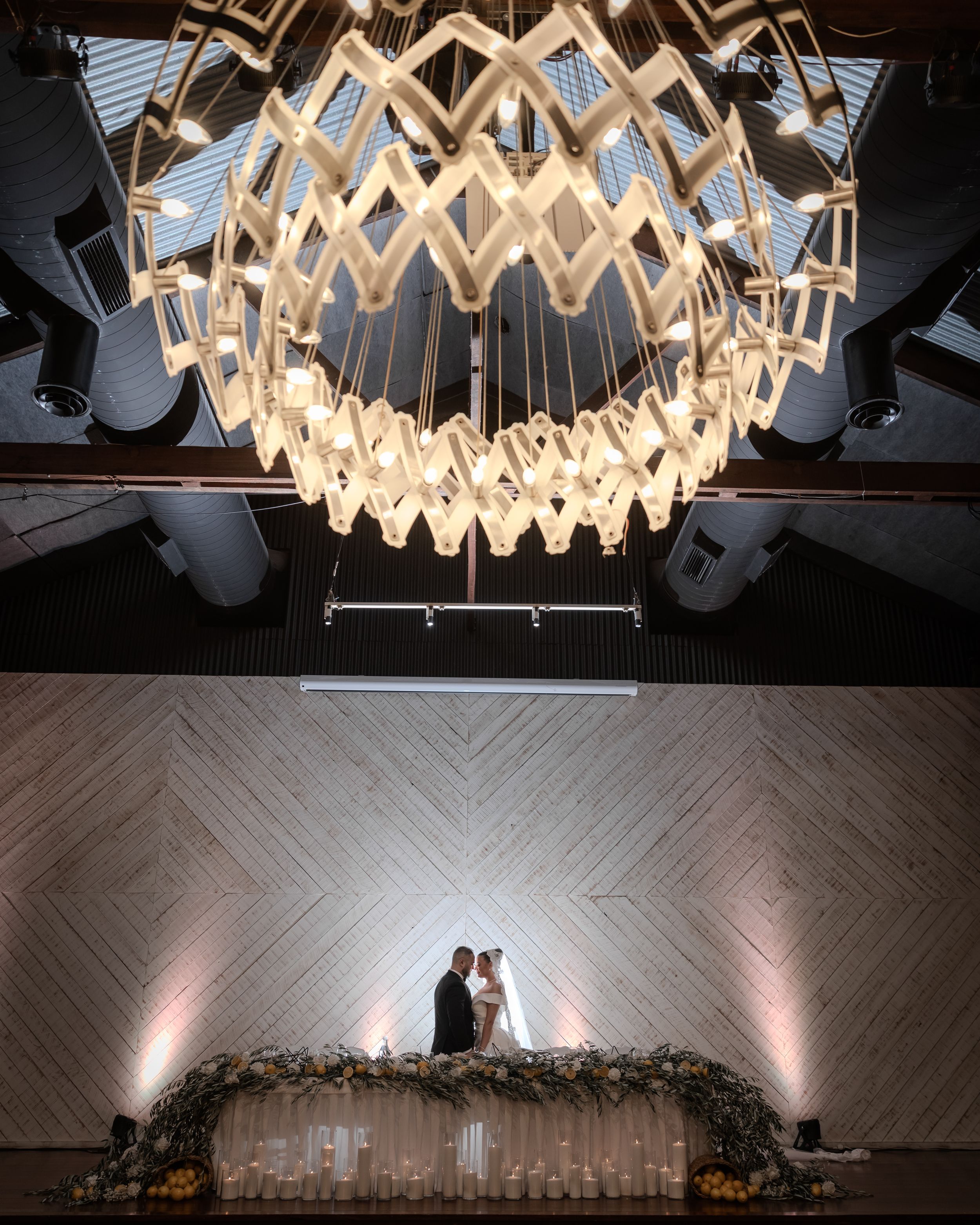 Couple shares intimate moment beneath an ornate geometric chandelier above a floral arch with candles.