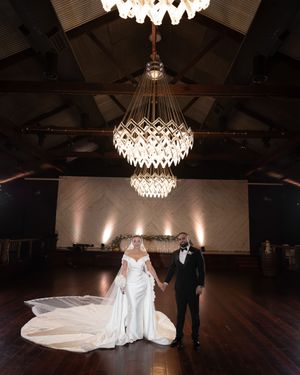 Bride and groom hold hands beneath dramatic geometric crystal chandeliers on a dark hardwood dance floor.