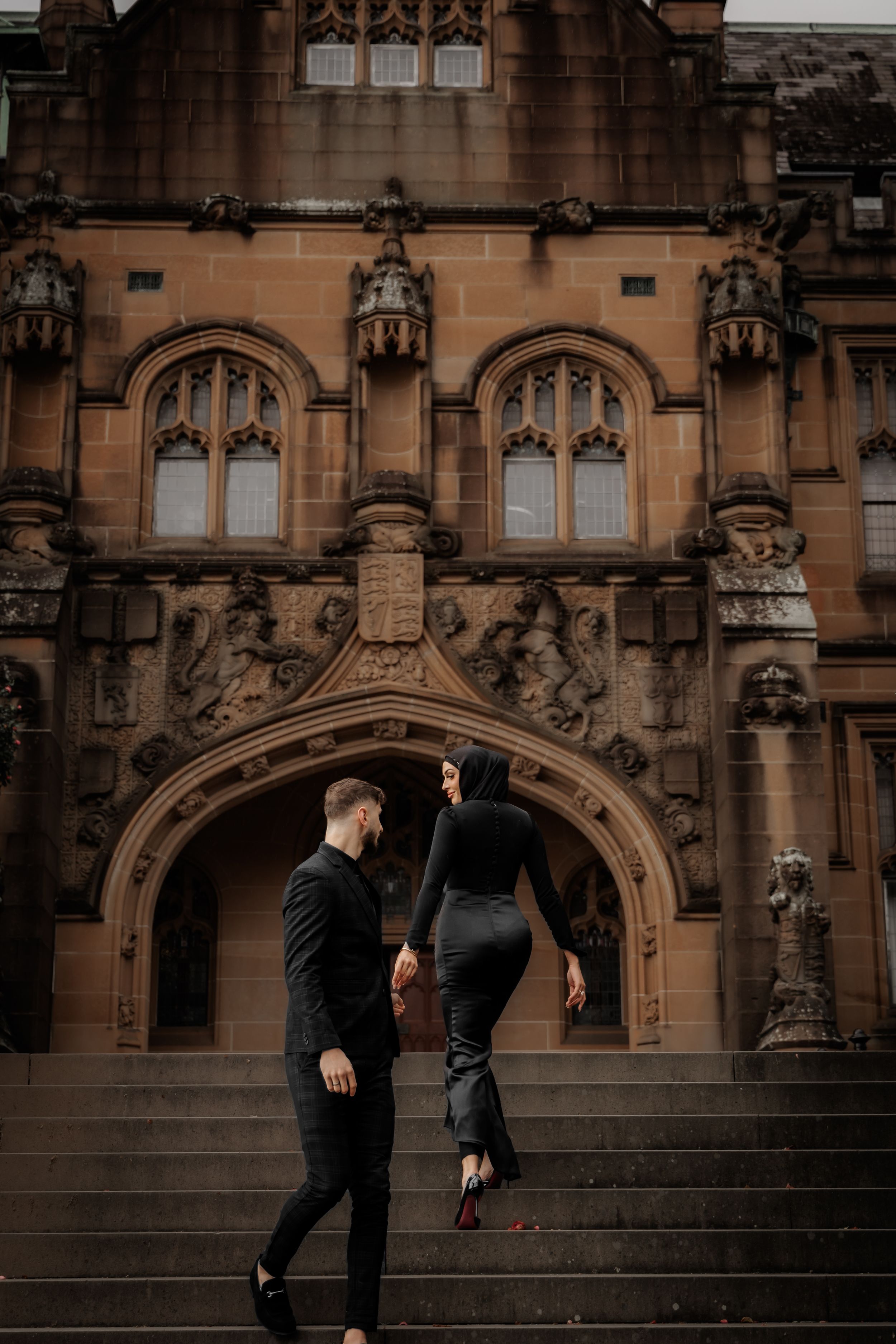 Stylish couple in all-black outfits pose on steps before ornate gothic sandstone building facade.